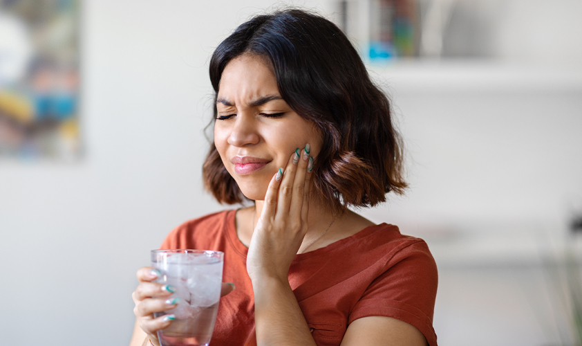 Woman wincing in pain from sensitive teeth while holding a glass of ice water, illustrating dental hypersensitivity symptoms.