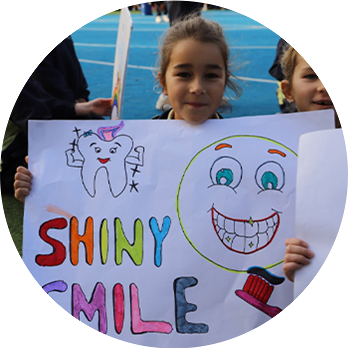A young girl holding up a colorful hand-drawn poster that says 'SHINY SMILE', featuring drawings of a cartoon tooth, a smiling face with clean teeth, and a toothbrush.